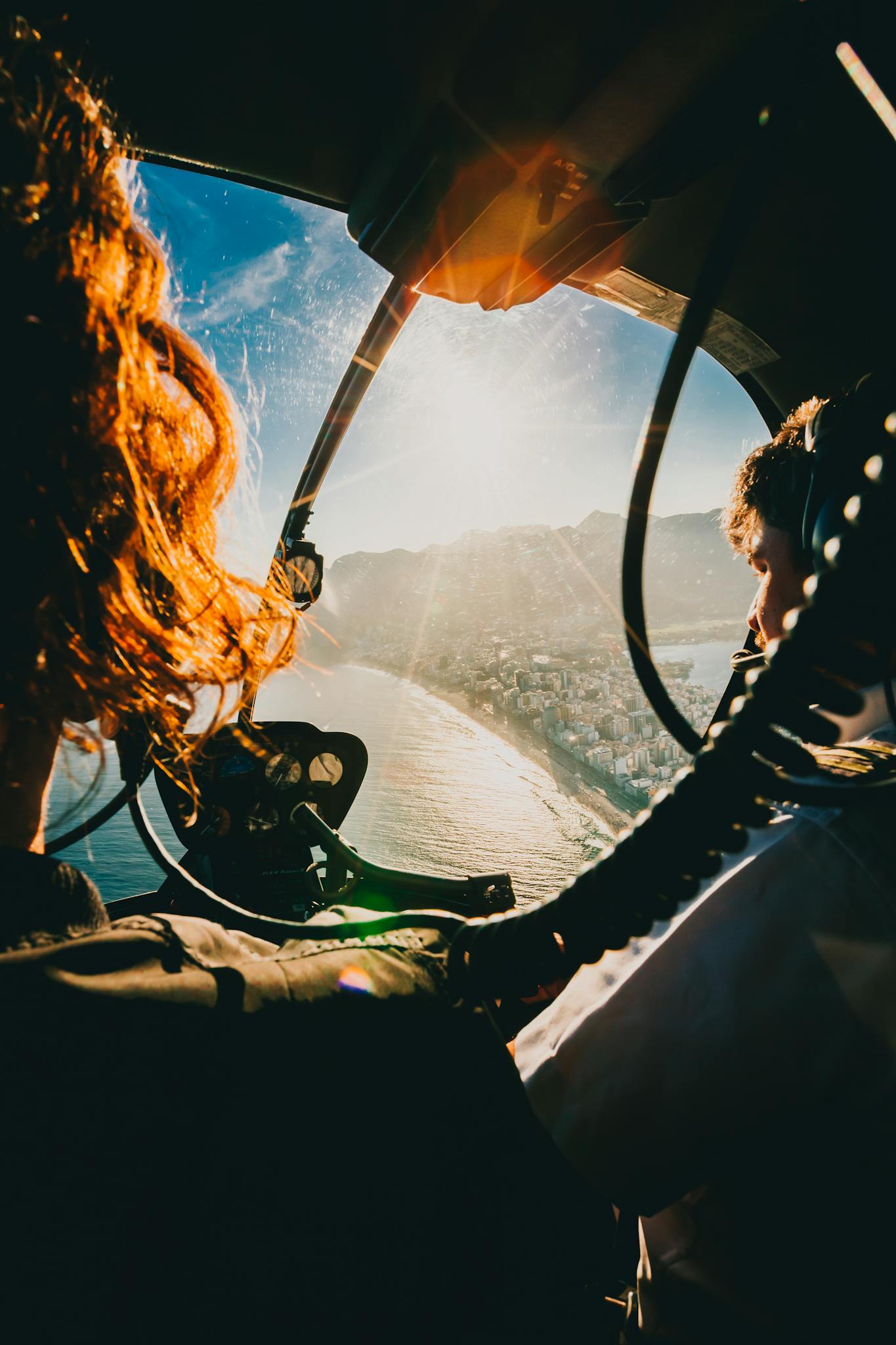 A pilot navigates a helicopter above the scenic coastline of Rio de Janeiro at sunrise.